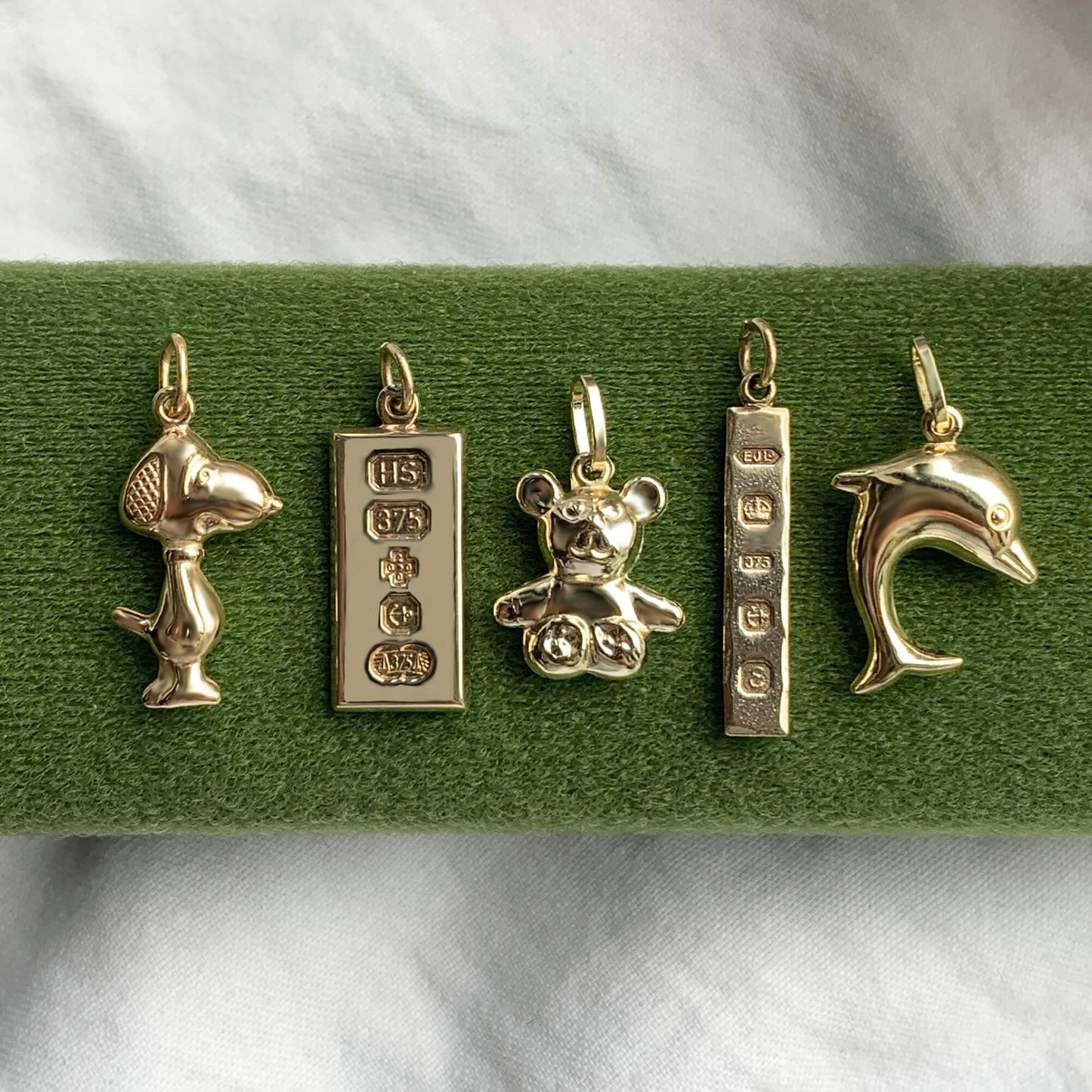 5 gold charms on a green jewellery box- left to right snoopy, millennium ingot, teddy bear, slim ingot, dolphin ingot.